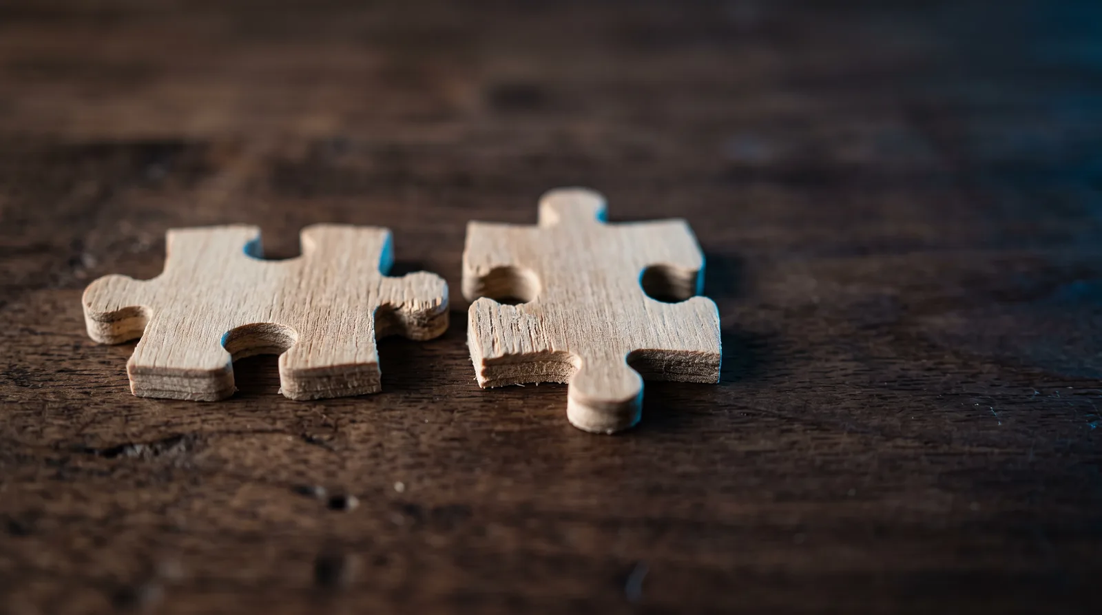 Two puzzle pieces that almost fit together sitting on a wooden table under soft blue light.