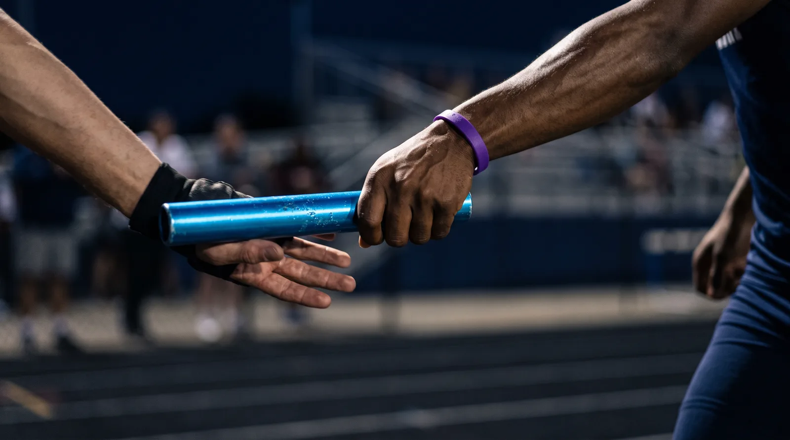 Two runners' hands pass a blue relay baton mid stride against a dark background, one wrist wearing a purple band.
