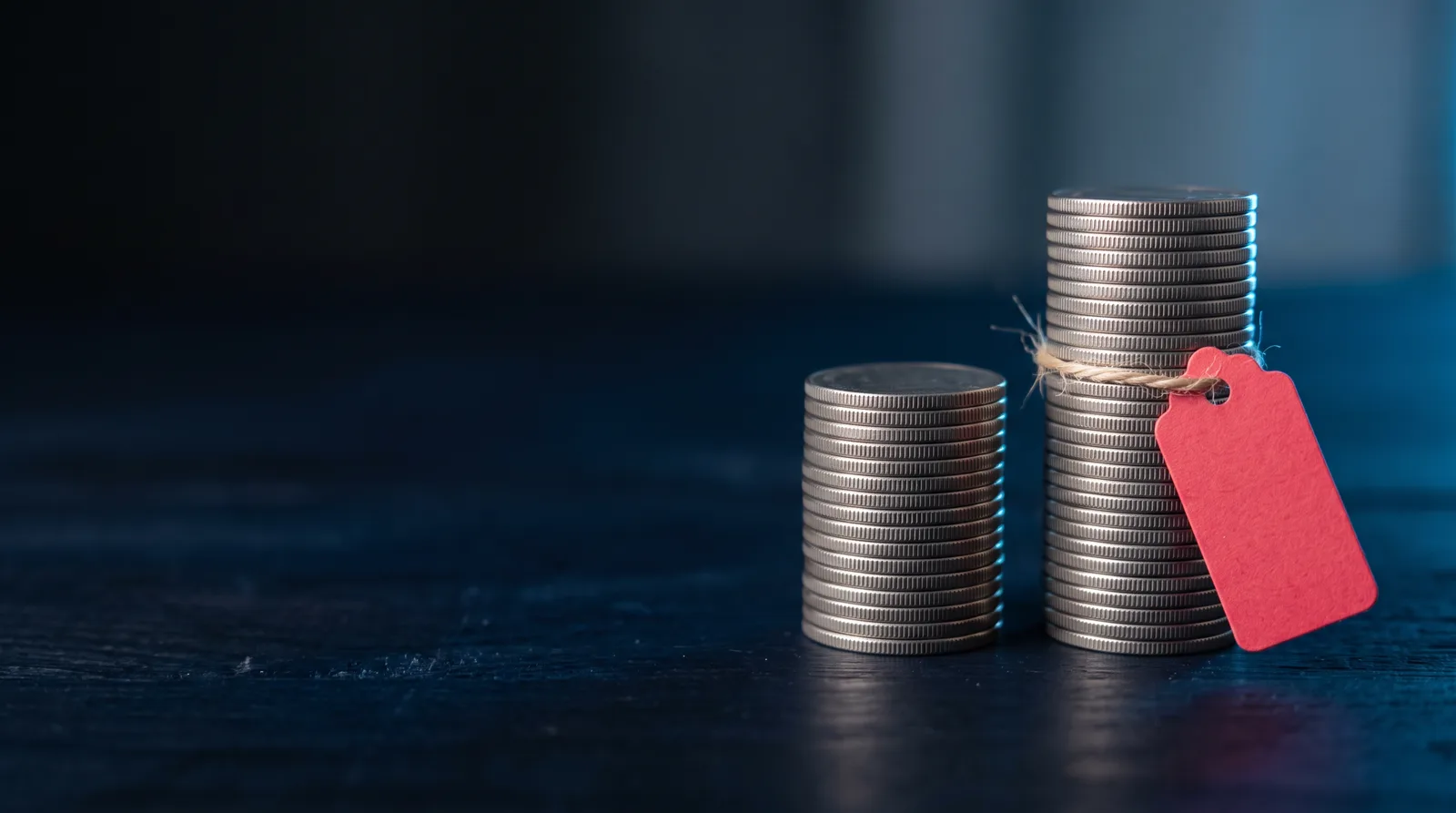 Two stacks of plain coins on a dark table, the right stack exactly twice the height of the left, with a small red tag tied to the taller stack.